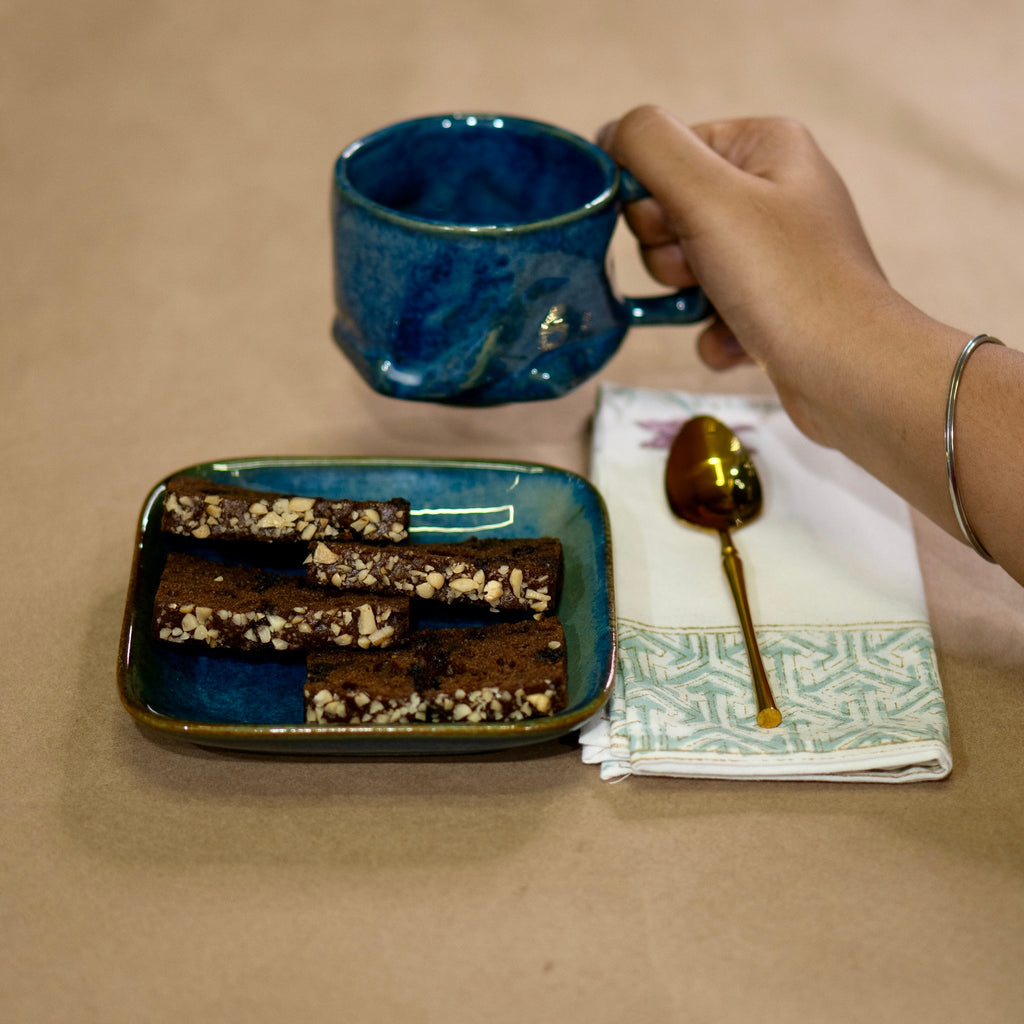 Hand holding a blue mug above a plate of brownies on a beige surface