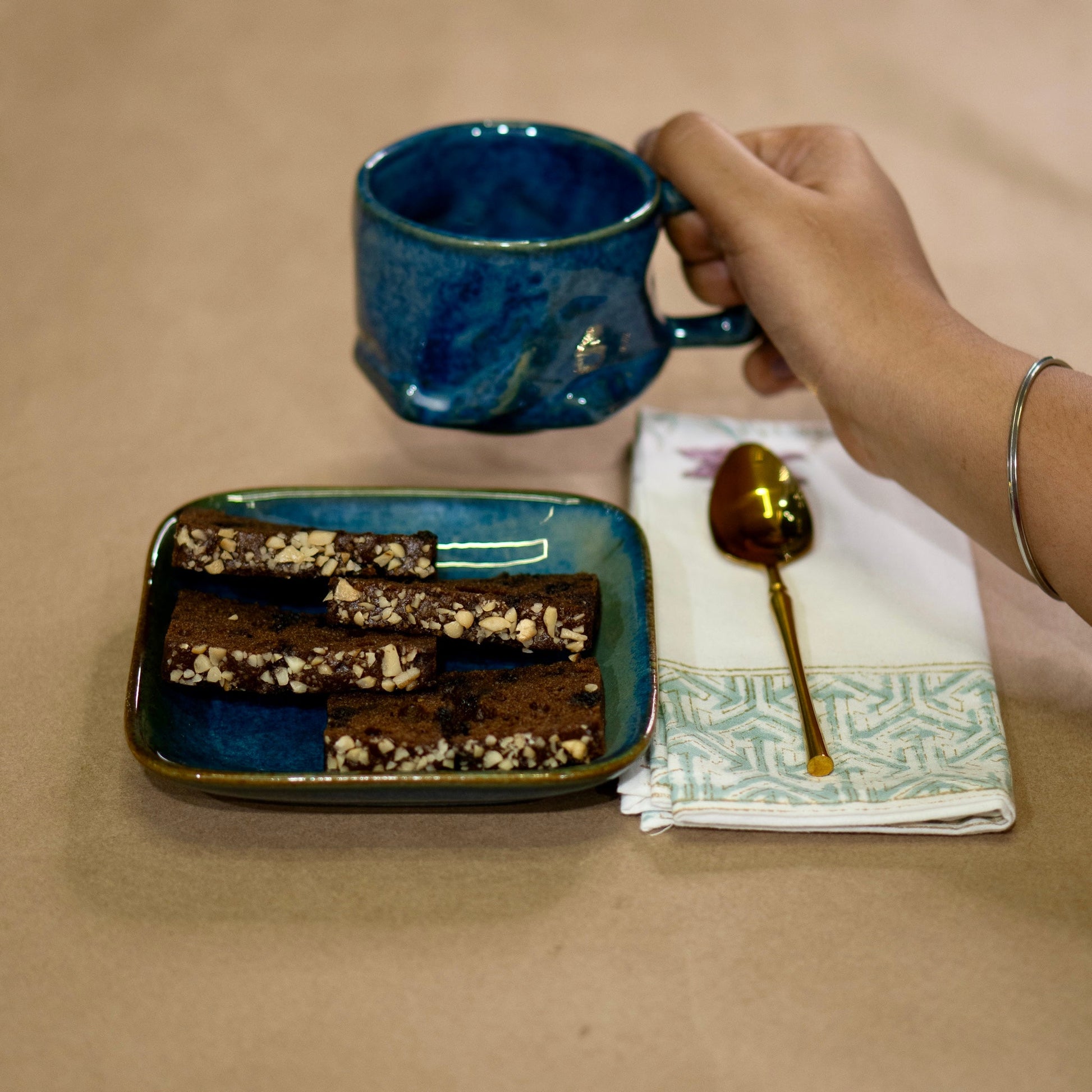 Hand holding a blue mug above a plate of brownies on a beige surface