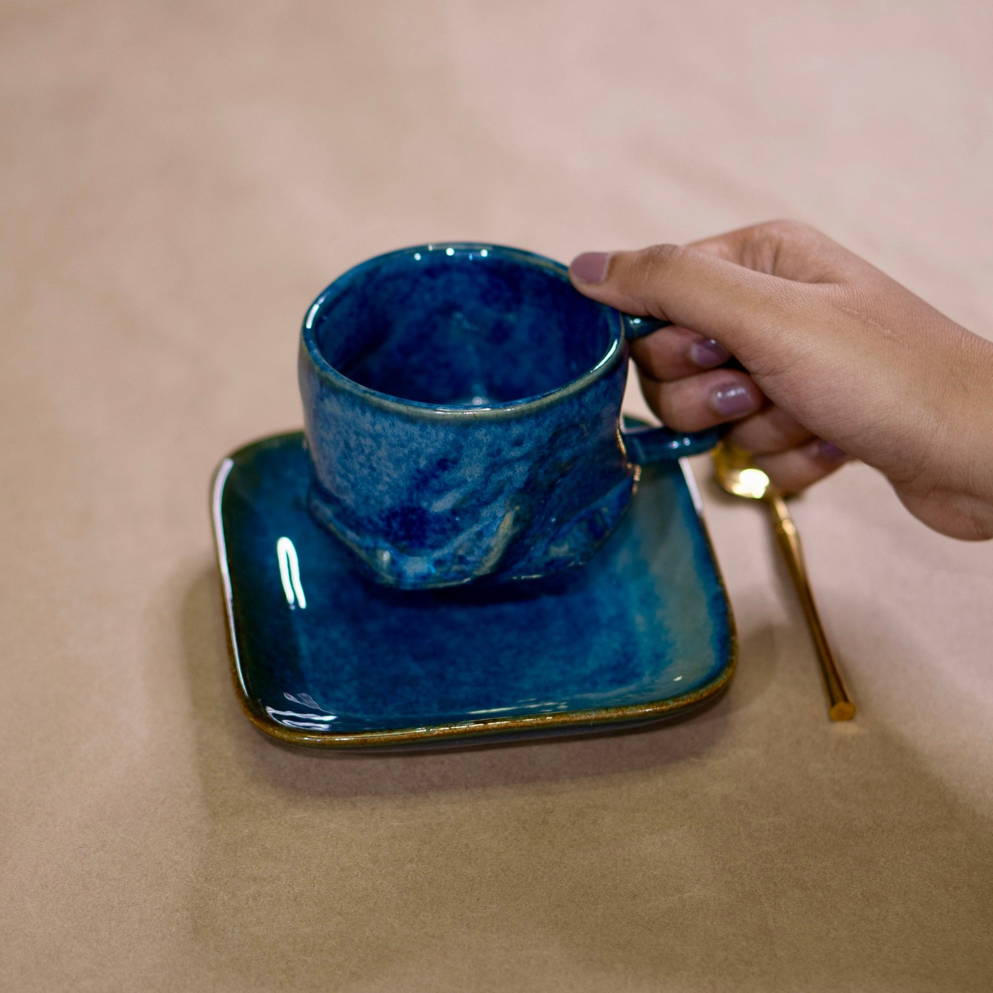 Hand holding a blue ceramic cup with saucer on a beige surface