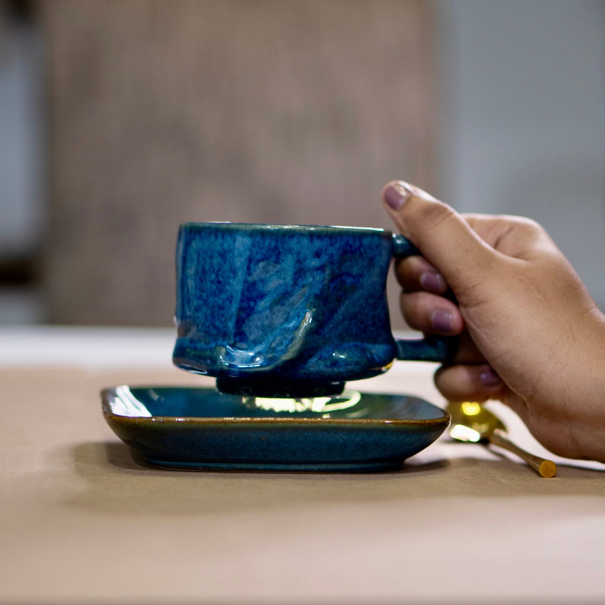 Hand holding a blue ceramic mug on a matching saucer with a blurred background