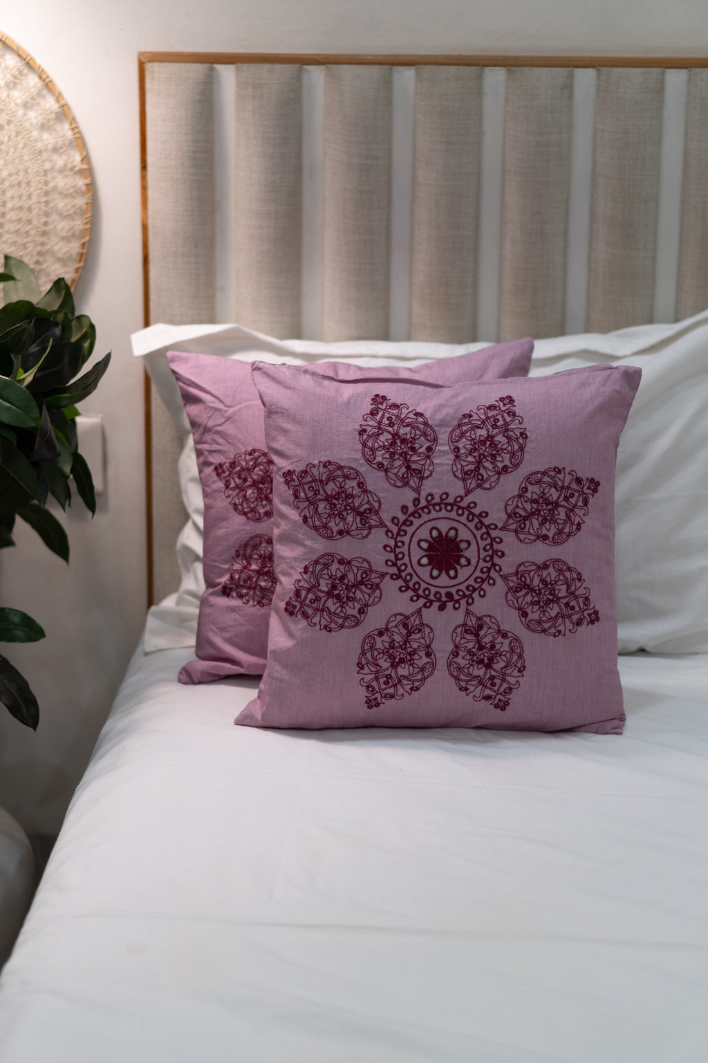 Pink decorative pillow with a floral pattern on a bed in a bedroom setting. Our Embroidery Cushion with Intricately embroidered mandala motifs. Schiffli Embroidered Cushions with cutwork.